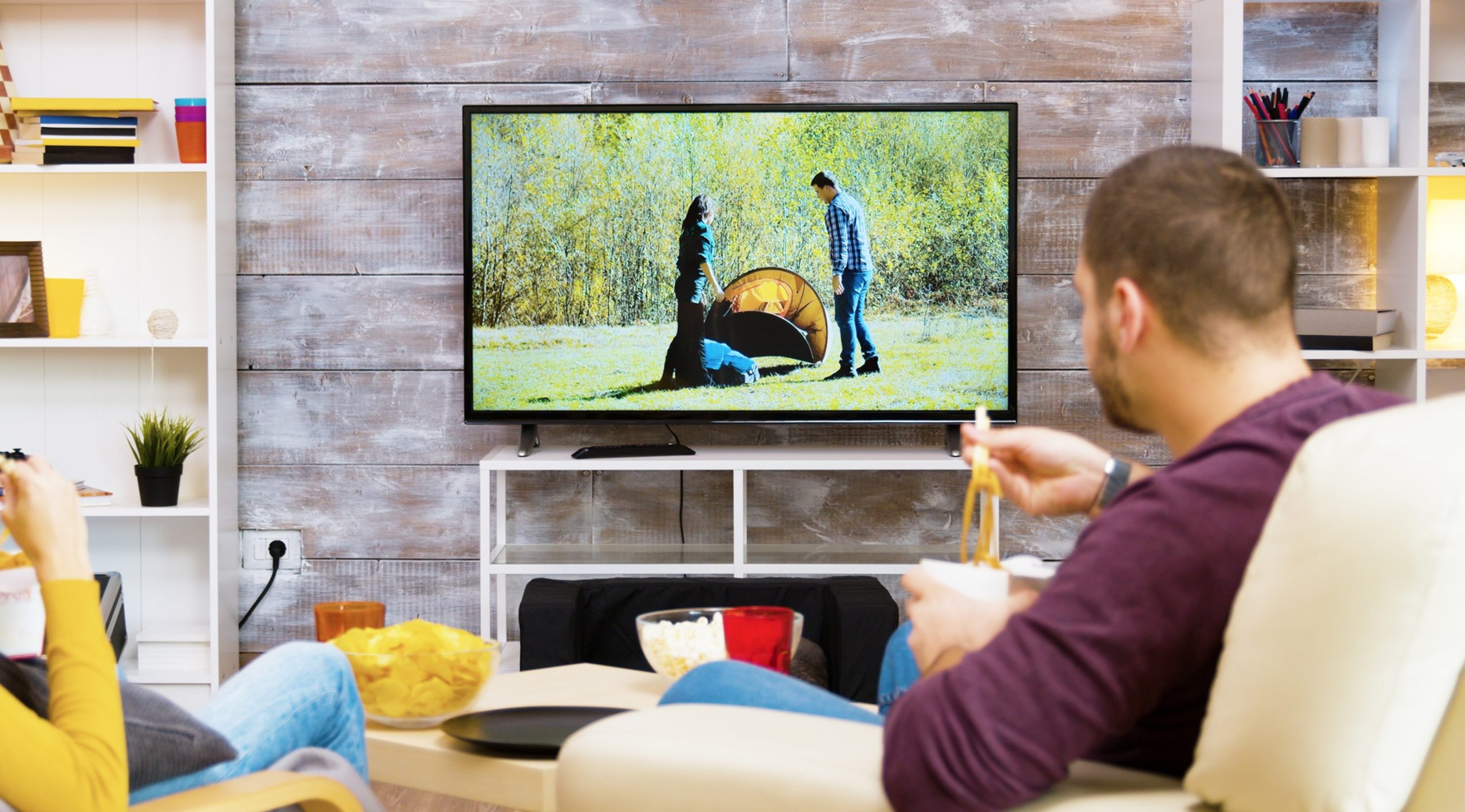 Two people sit on a couch, eating snacks while watching a camping scene and listening to a commercial voice over on a flat-screen TV in their living room.