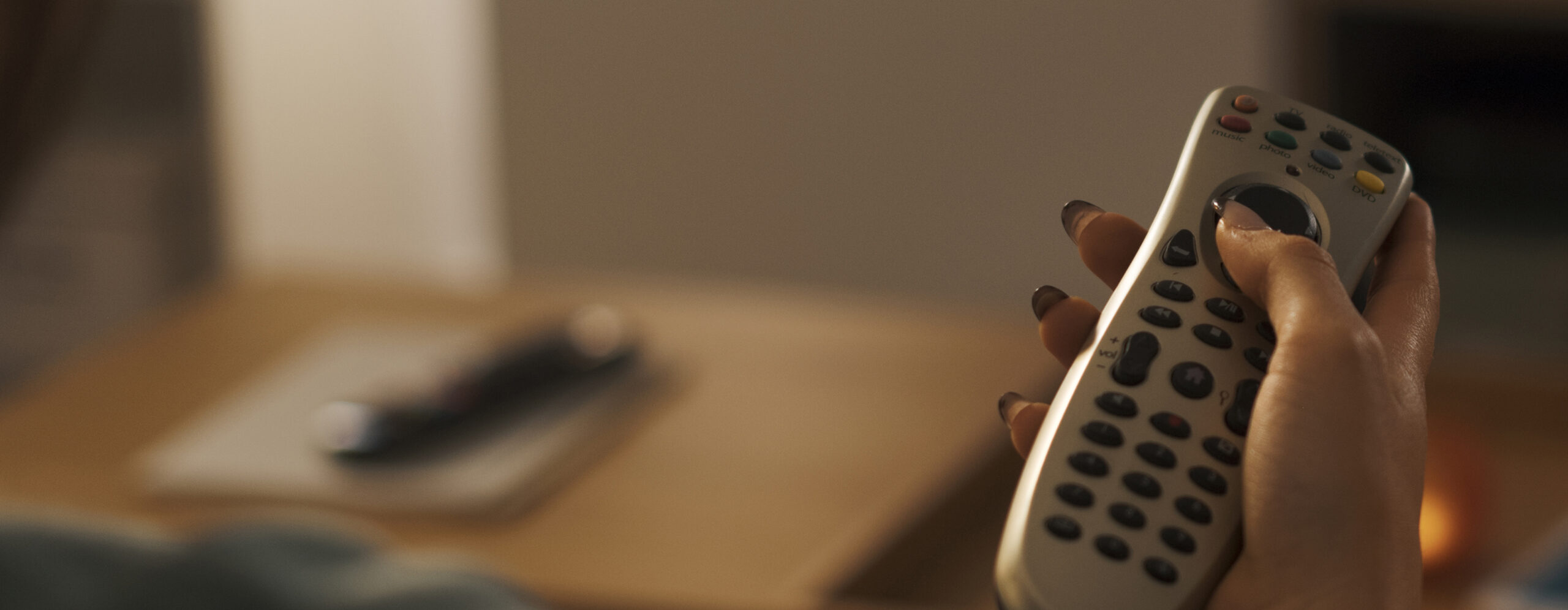 A person holds and uses a television remote control—perhaps searching for the perfect channel to enjoy a commercial voice over—while sitting near a table with another remote on it.