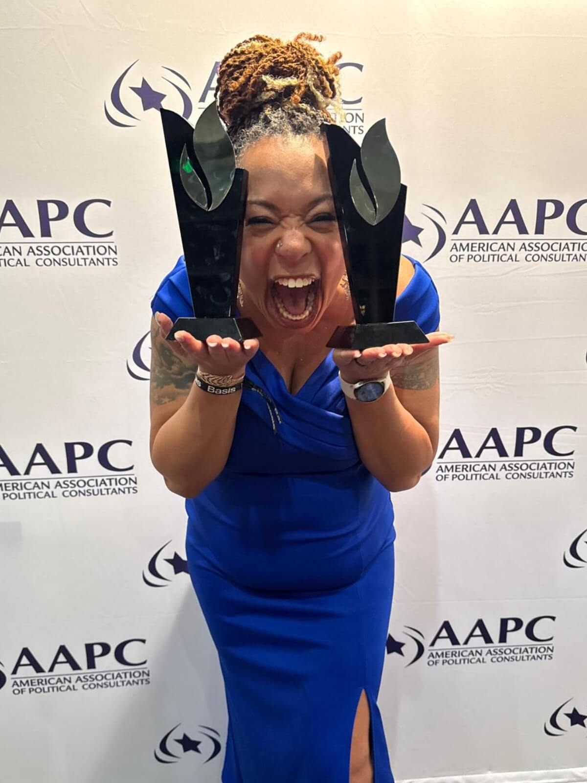 A woman in a blue dress holds two black awards and smiles widely in front of an American Association of Political Consultants step-and-repeat banner, celebrating her achievements in political voice over.
