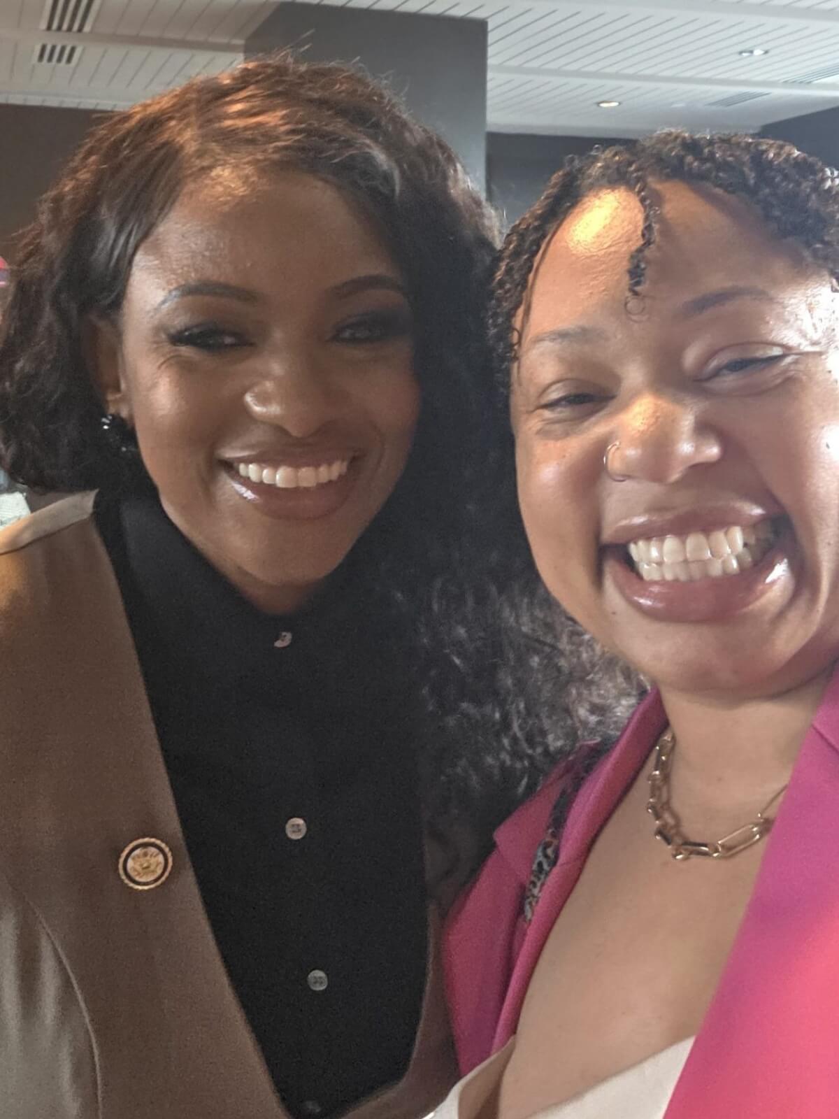 Two women smiling at the camera indoors, one wearing a brown blazer with a pin and the other in a pink blazer and necklace, as if preparing for a political voice over.