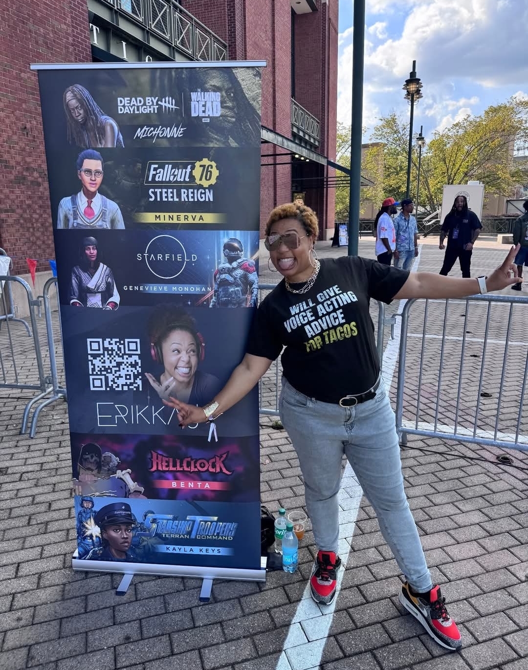 A person stands outdoors next to a vertical banner displaying promotional posters for video games and voice actors, smiling and gesturing towards the banner that highlights exciting video game voice over talent.