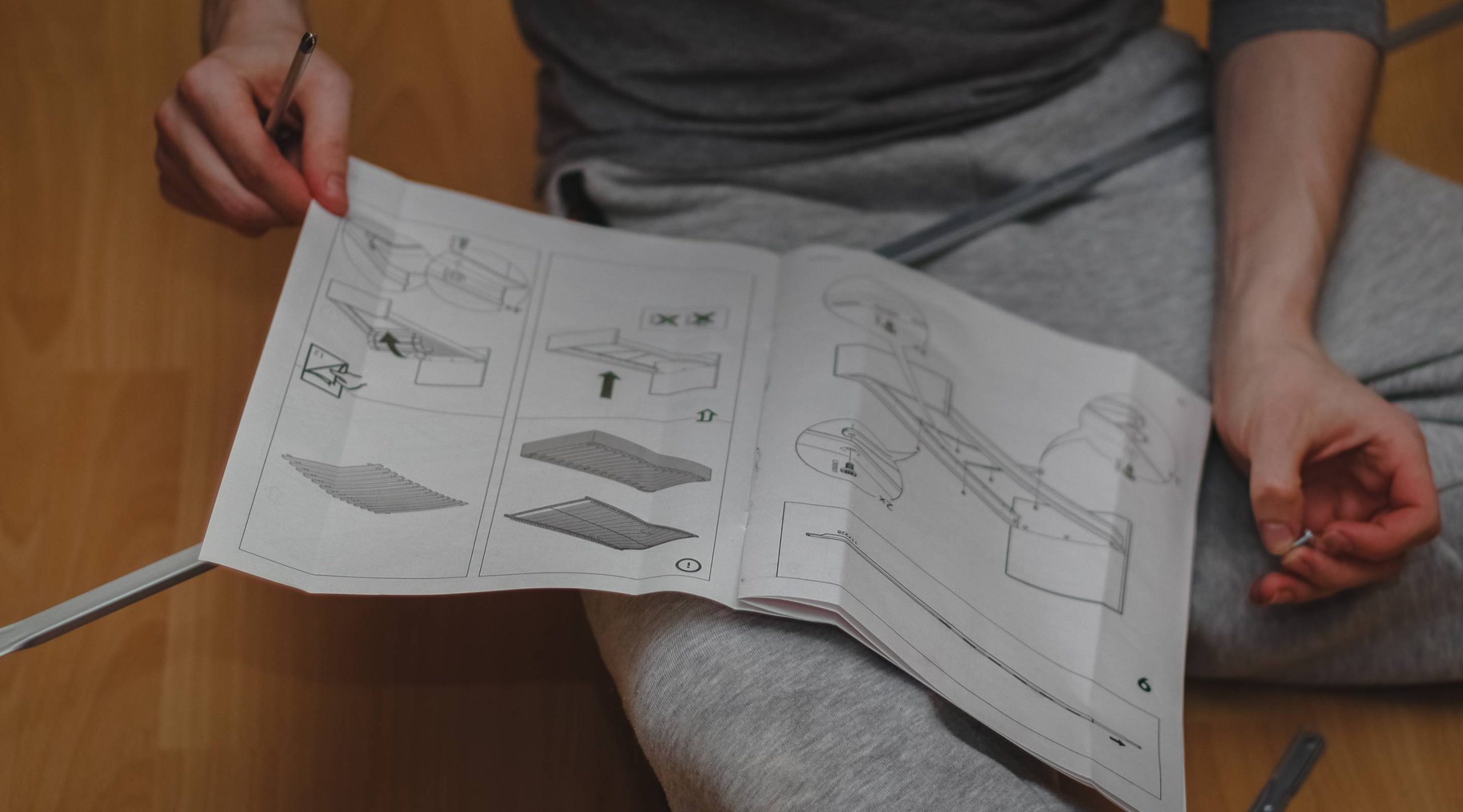 Person in grey clothing holding and looking at illustrated furniture assembly instructions while sitting on a wooden floor.
