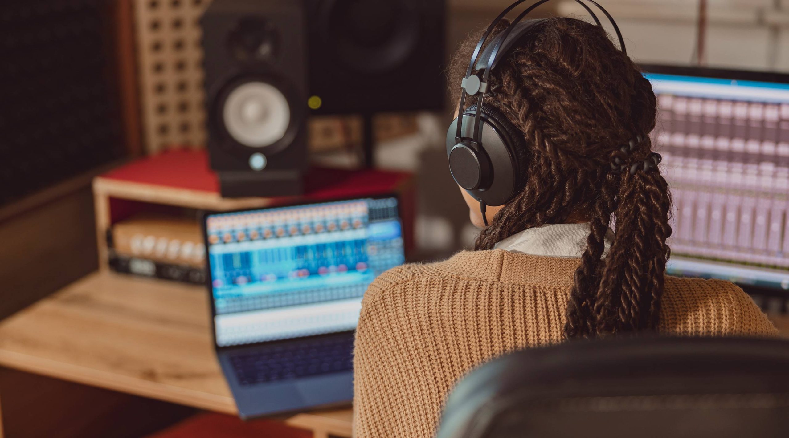 Person with headphones sits at a desk using a laptop and audio equipment in a music production studio.