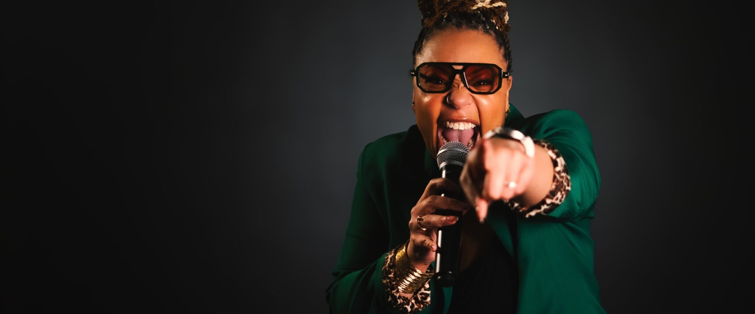Person wearing glasses and a green blazer holds a microphone, striking a broadcast-ready pose while pointing toward the camera against a dark background.
