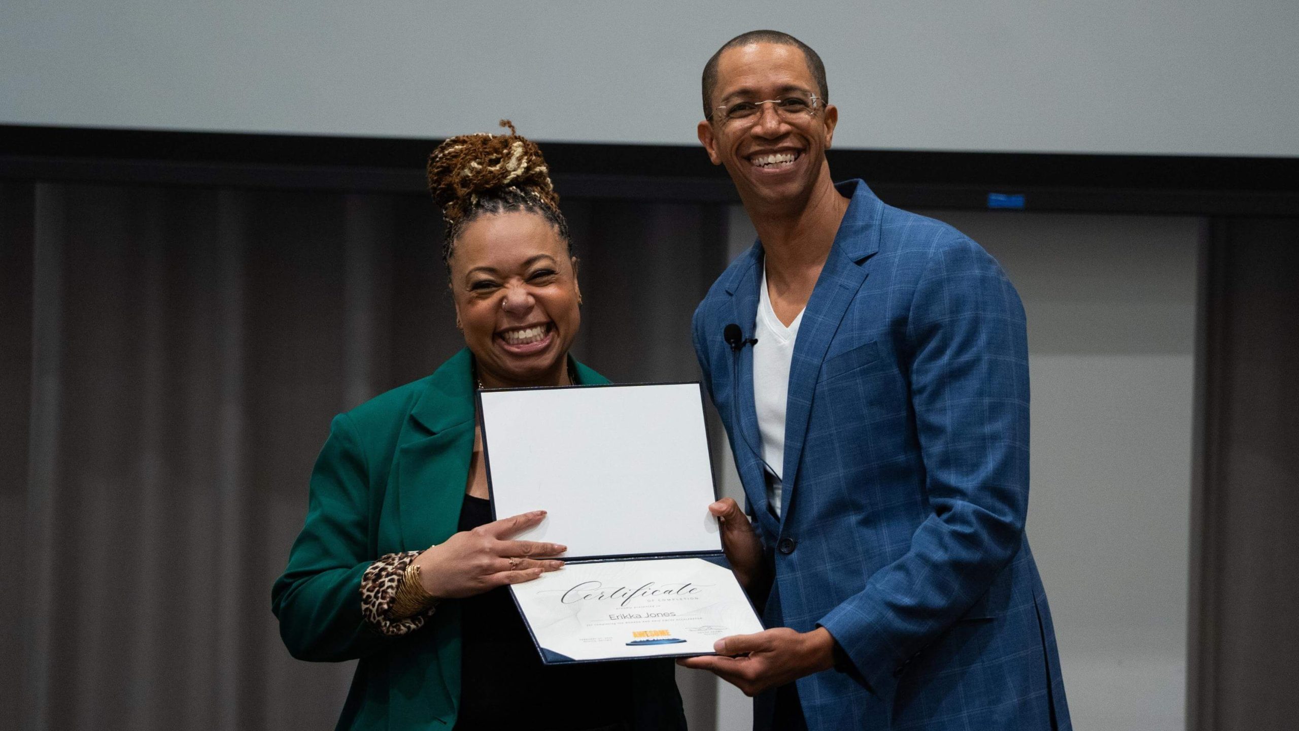 Erikka J and Devon smiling; one is holding an open certificate folder while posing for a photo at an indoor broadcast event.