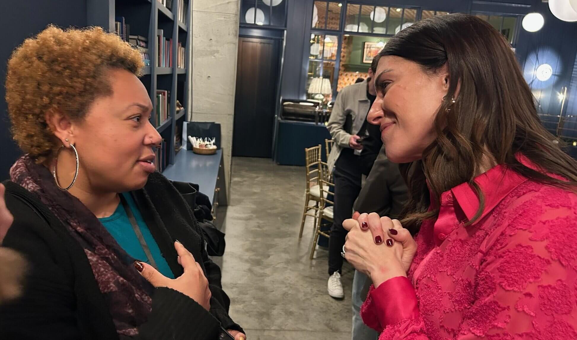 Two women stand facing each other indoors, engaged in conversation. One has curly hair and wears hoop earrings, while the other, dressed in a bright pink blouse, discusses details about a corporate voice over project.