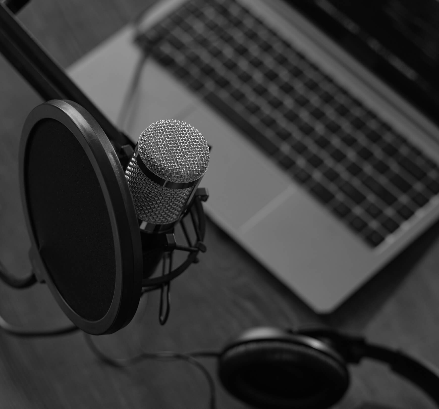 A microphone with a pop filter, a laptop, and headphones are arranged on a wooden surface, suggesting a recording or podcast setup.