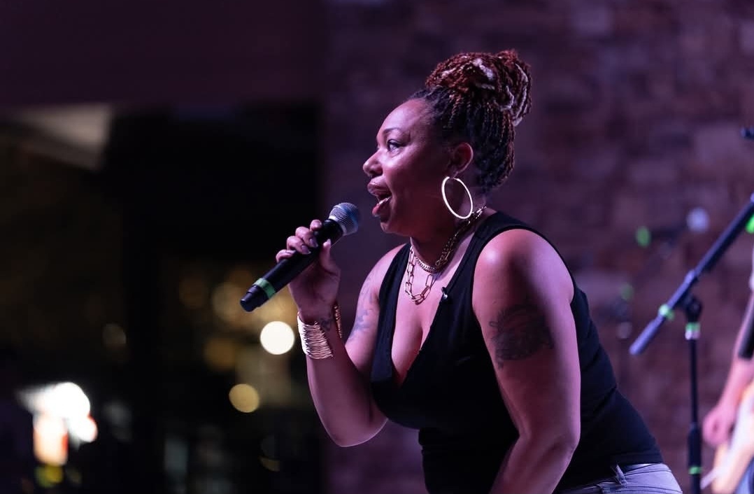 A woman with braided hair sings into a microphone on stage, wearing a black sleeveless top and hoop earrings, as her live broadcast captivates viewers against a blurred background.