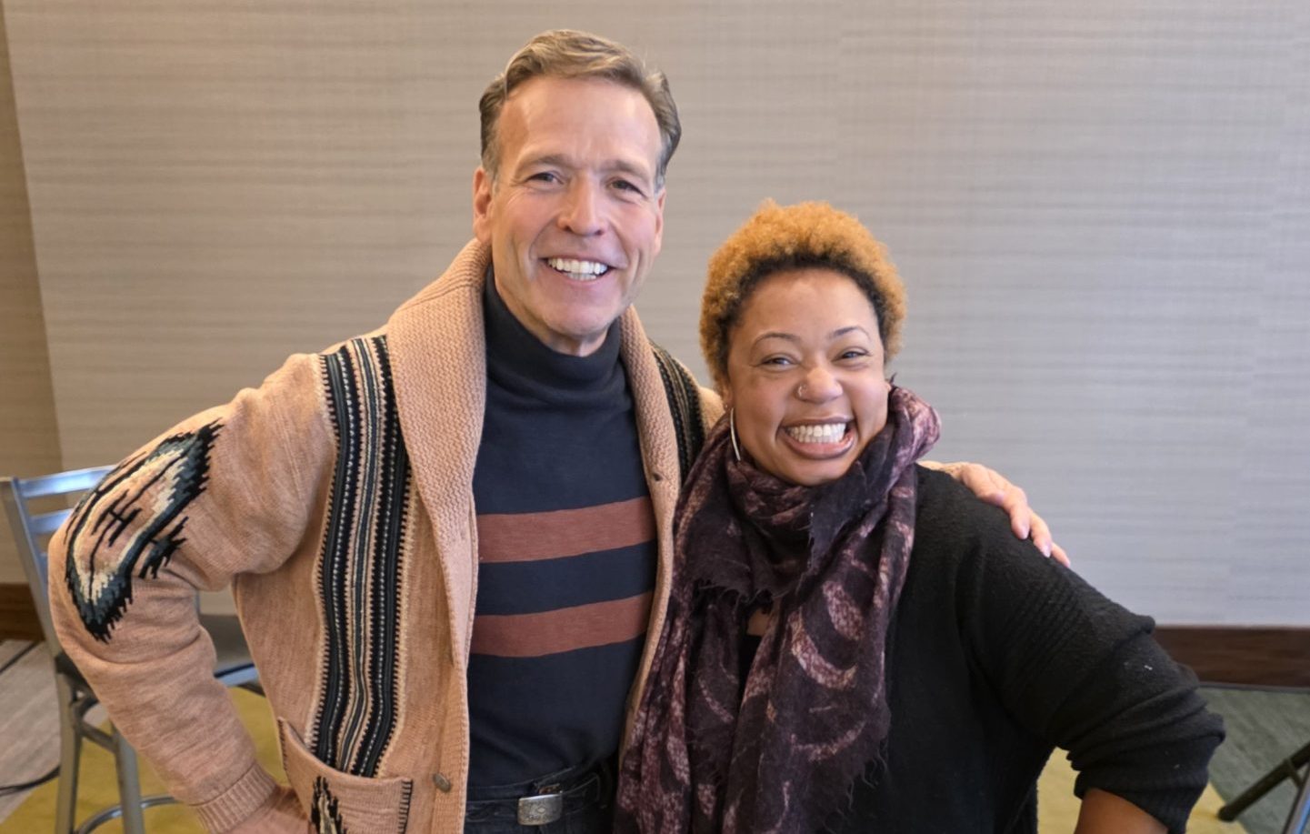 Two people stand side by side indoors, smiling at the camera. The person on the left wears jeans, a striped shirt, and a tan sweater; the person on the right, perfect for a corporate voice over project, wears a black outfit with a patterned scarf.