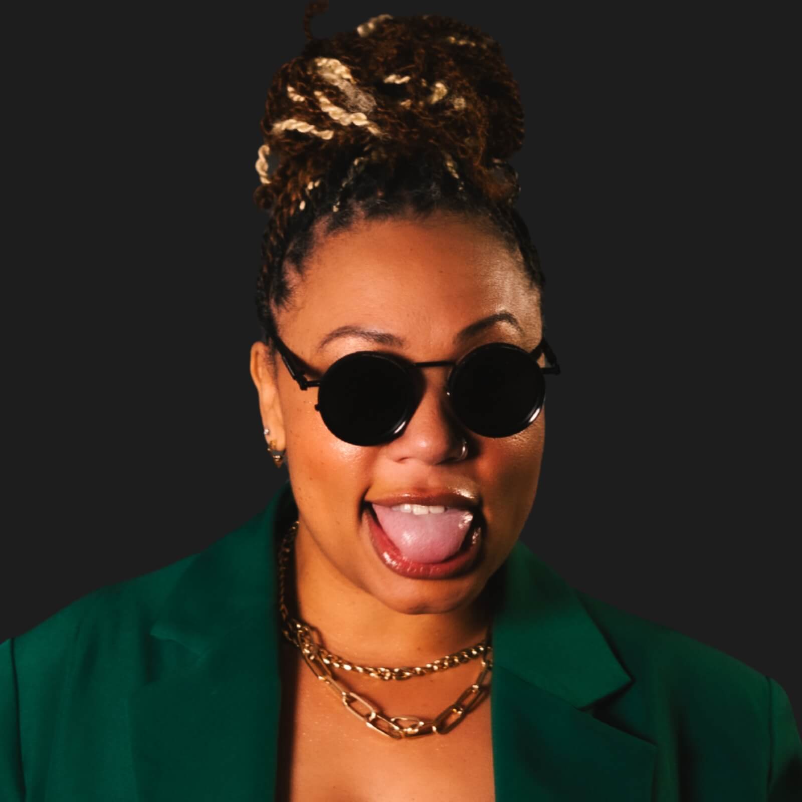 A person wearing round black sunglasses, gold chains, and a green jacket sticks out their tongue against a plain dark background, channeling the playful energy of voice actor Erikka J.