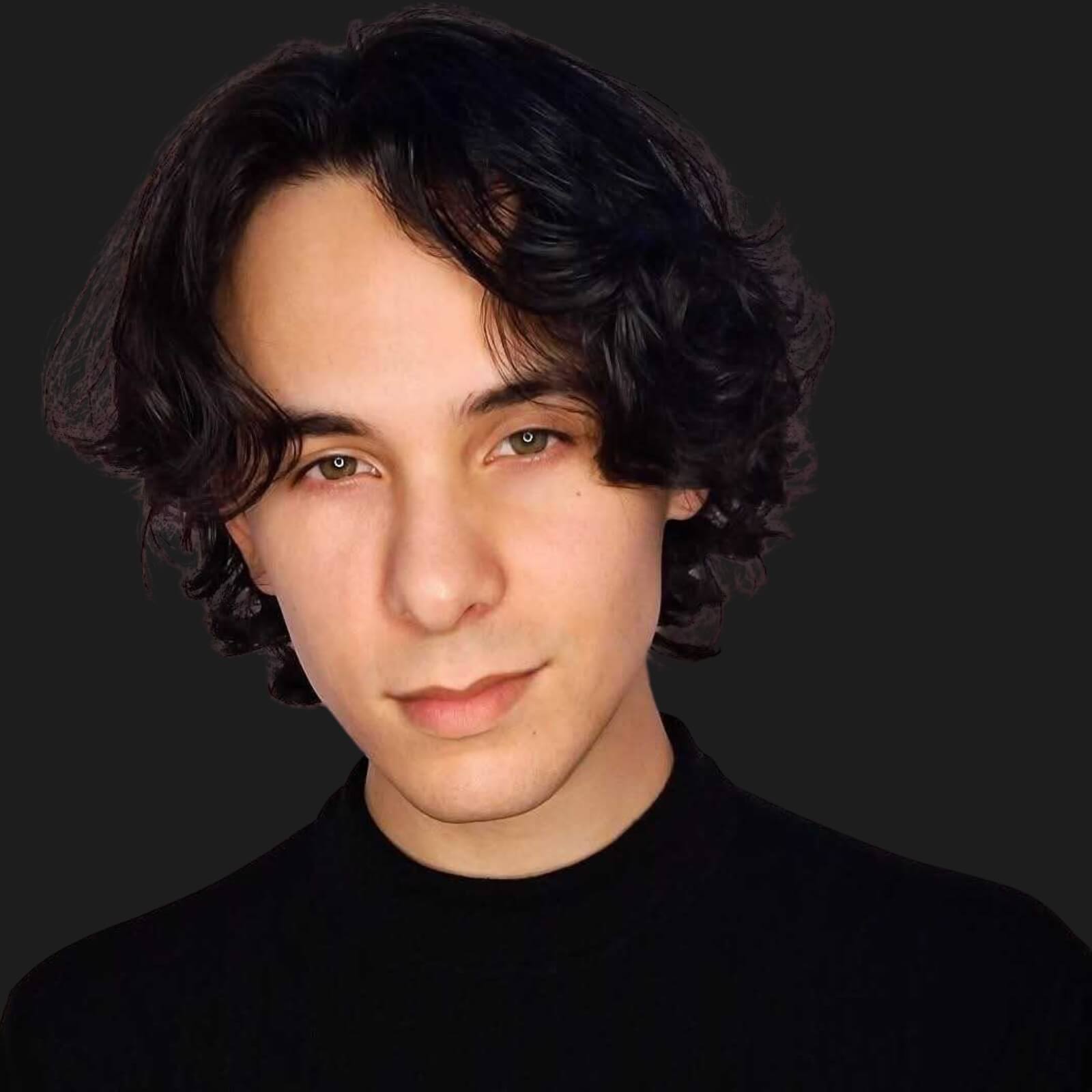 Young person with wavy, medium-length dark hair and fair skin, wearing a black turtleneck, faces forward against a plain dark background—reminiscent of voice actor Erikka J’s poised style.