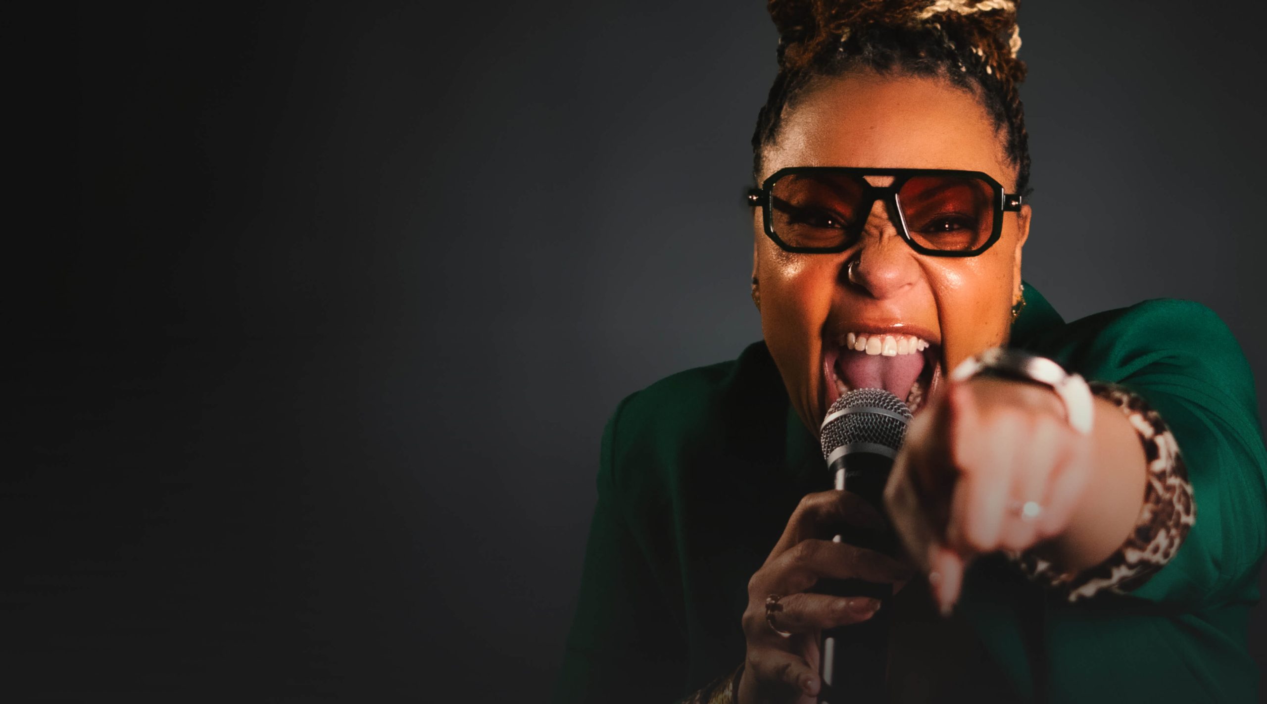 A person wearing sunglasses passionately sings into a microphone while pointing towards the camera, channeling energy reminiscent of voice actor Erikka J, against a dark background.