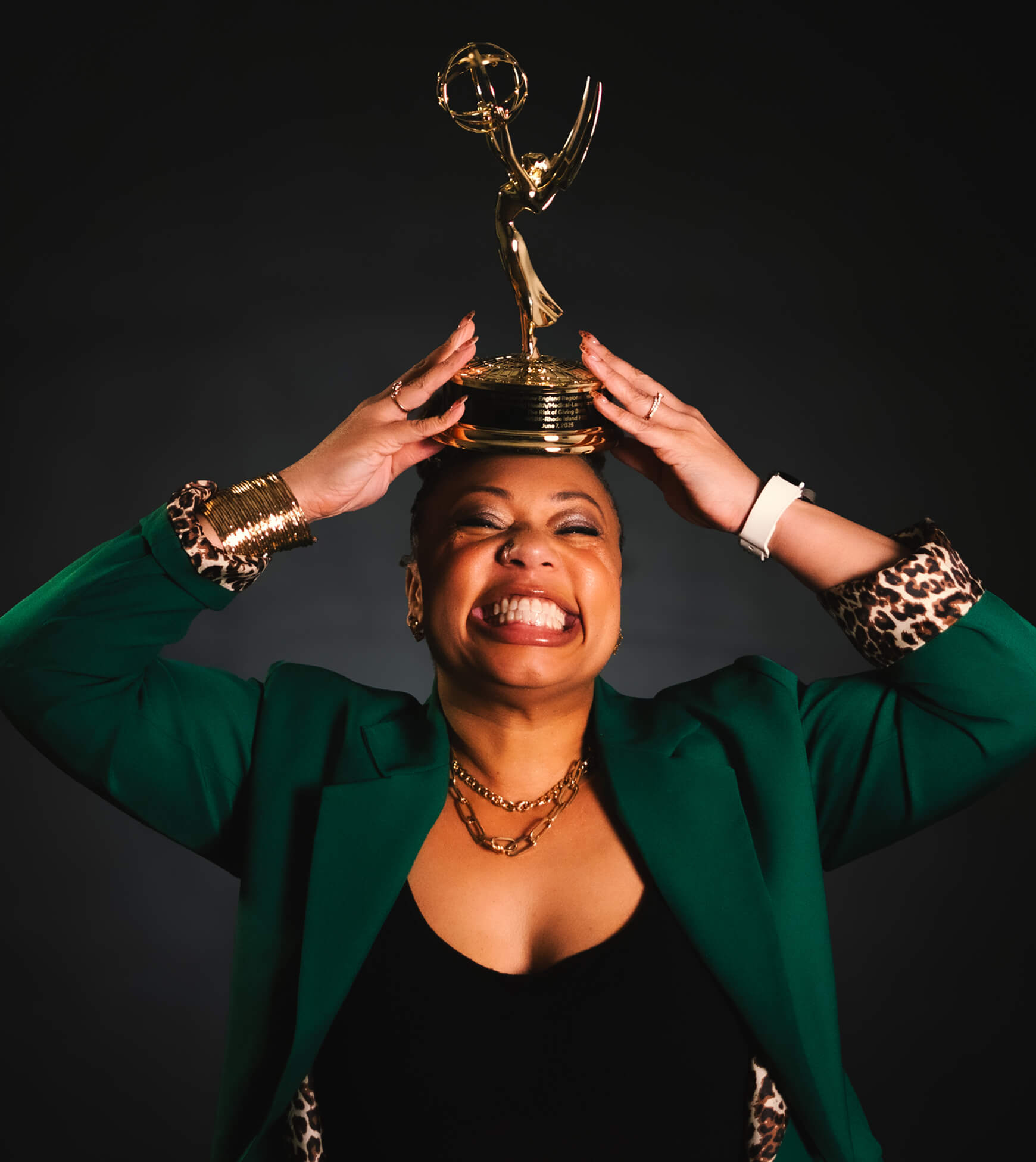 A person in a green jacket smiles broadly while holding an Emmy Award trophy above their head against a dark background, celebrating voice actor Erikka J's remarkable achievement.