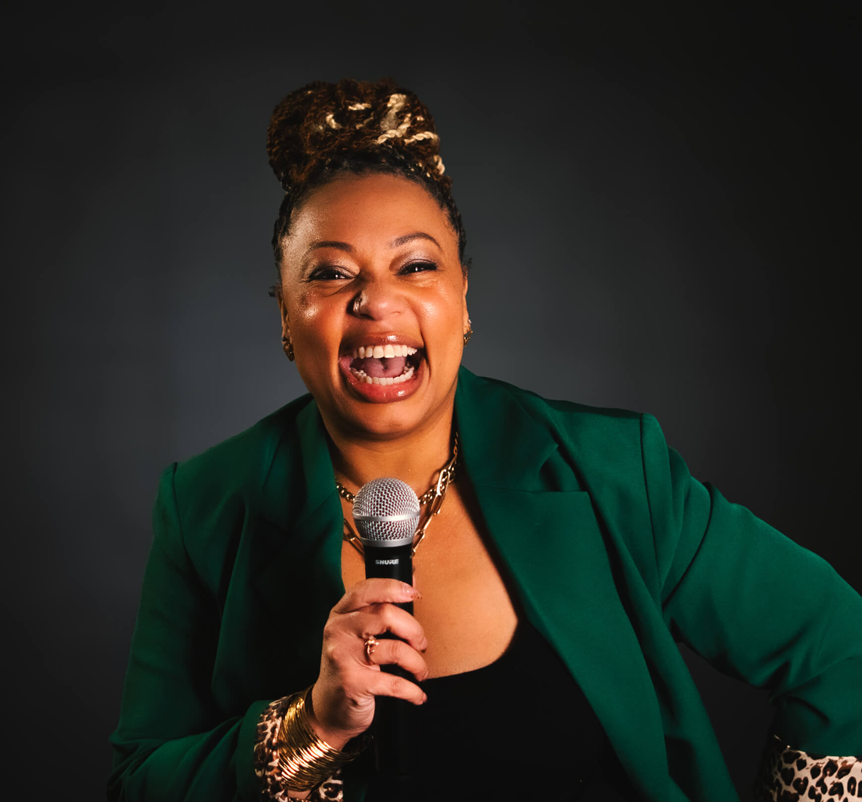 Woman wearing a green blazer and gold jewelry smiles widely while holding a microphone against a dark background, capturing the vibrant energy of voice actor Erikka J.