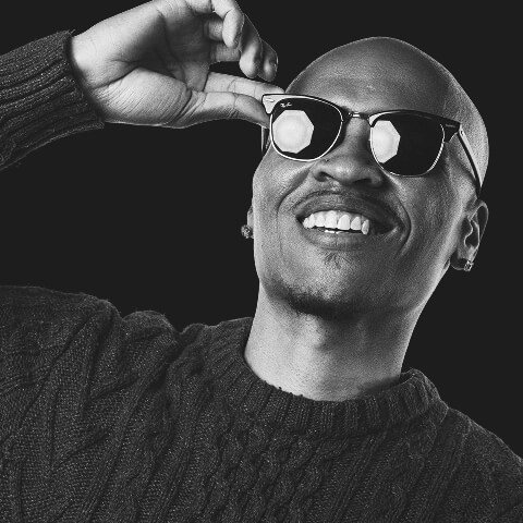 A person wearing sunglasses and a textured sweater smiles, tilting their head slightly back against a dark background—capturing the laid-back charisma of voice actor Erikka J.