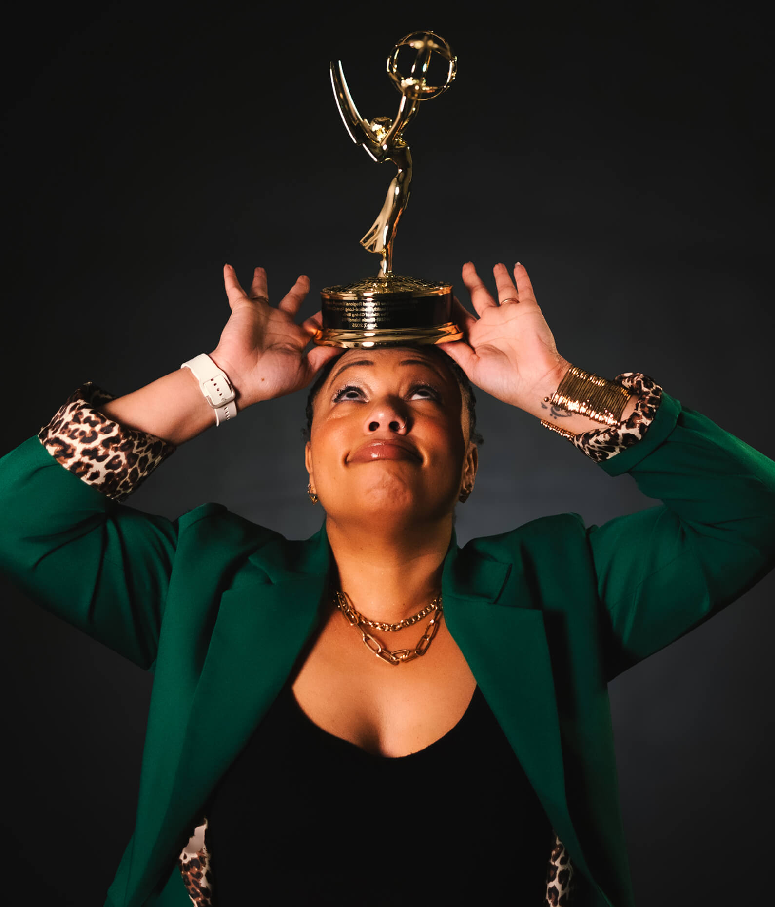 A voice actor in a green blazer balances an Emmy award on their head against a dark background, looking upward and holding the trophy with both hands.