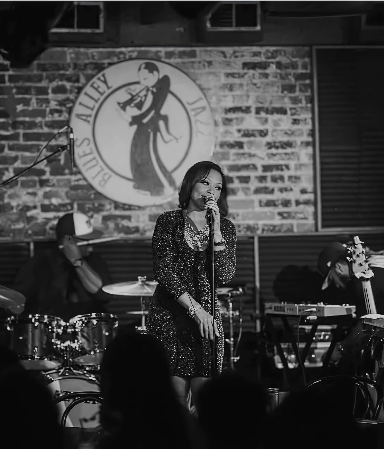 A woman sings on stage at a jazz club, with a band playing instruments behind her. The background features a sign reading "Blues Alley Jazz," and the soulful performance recalls the talent of voice actor Erikka J.