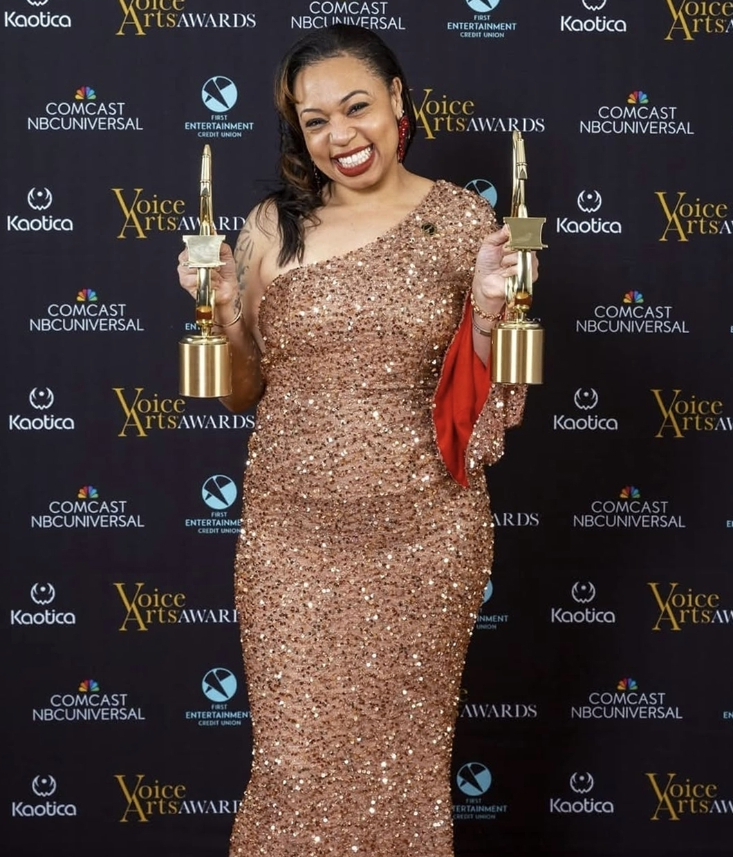 A woman in a sparkling one-shoulder gown, voice actor Erikka J, smiles and holds two trophies on the red carpet at the Voice Arts Awards, in front of a step-and-repeat backdrop.