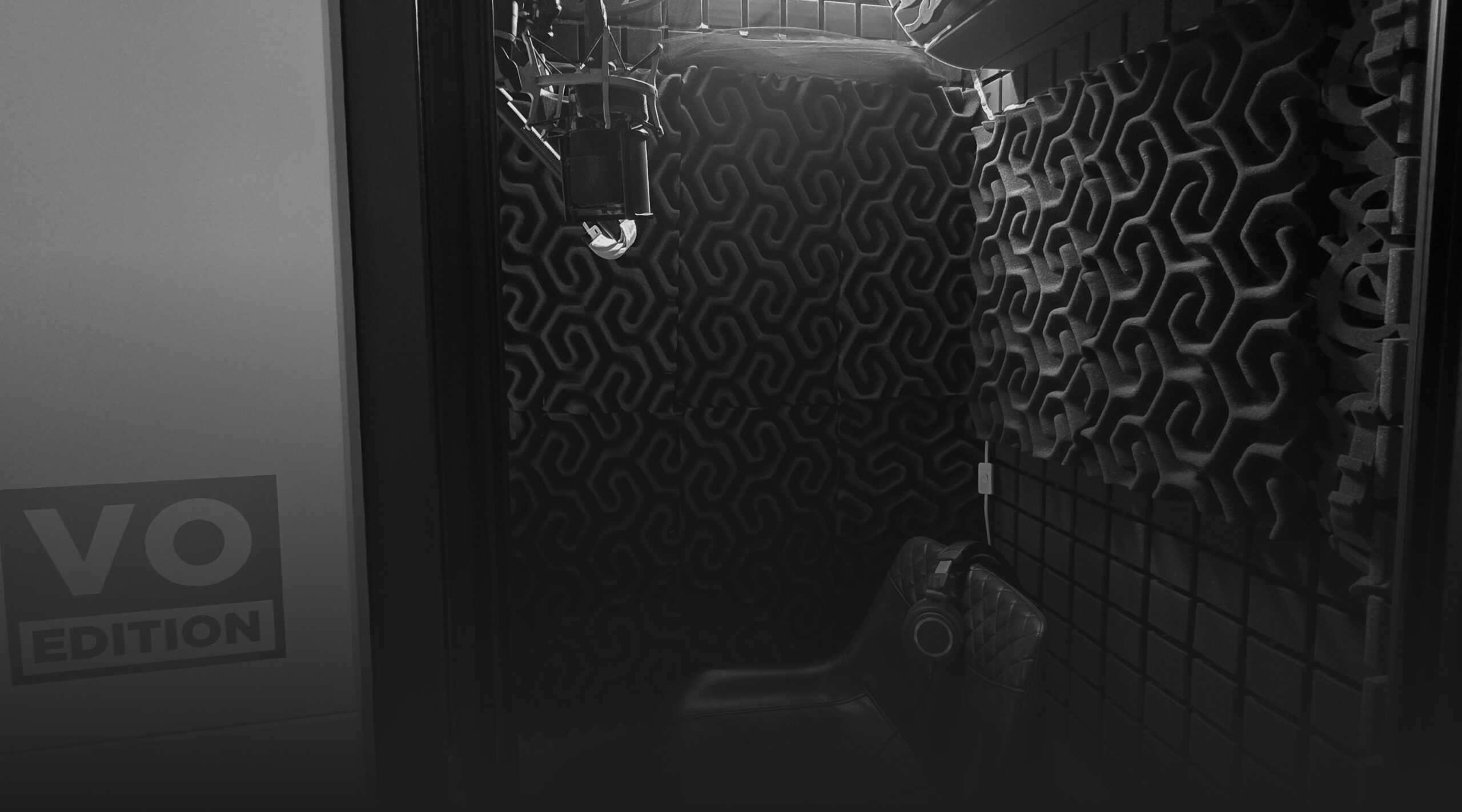 A small, soundproofed recording booth with foam-padded walls, a microphone, a chair, and a sign reading "VO Edition" on the door—perfect for capturing professional voiceover sessions.