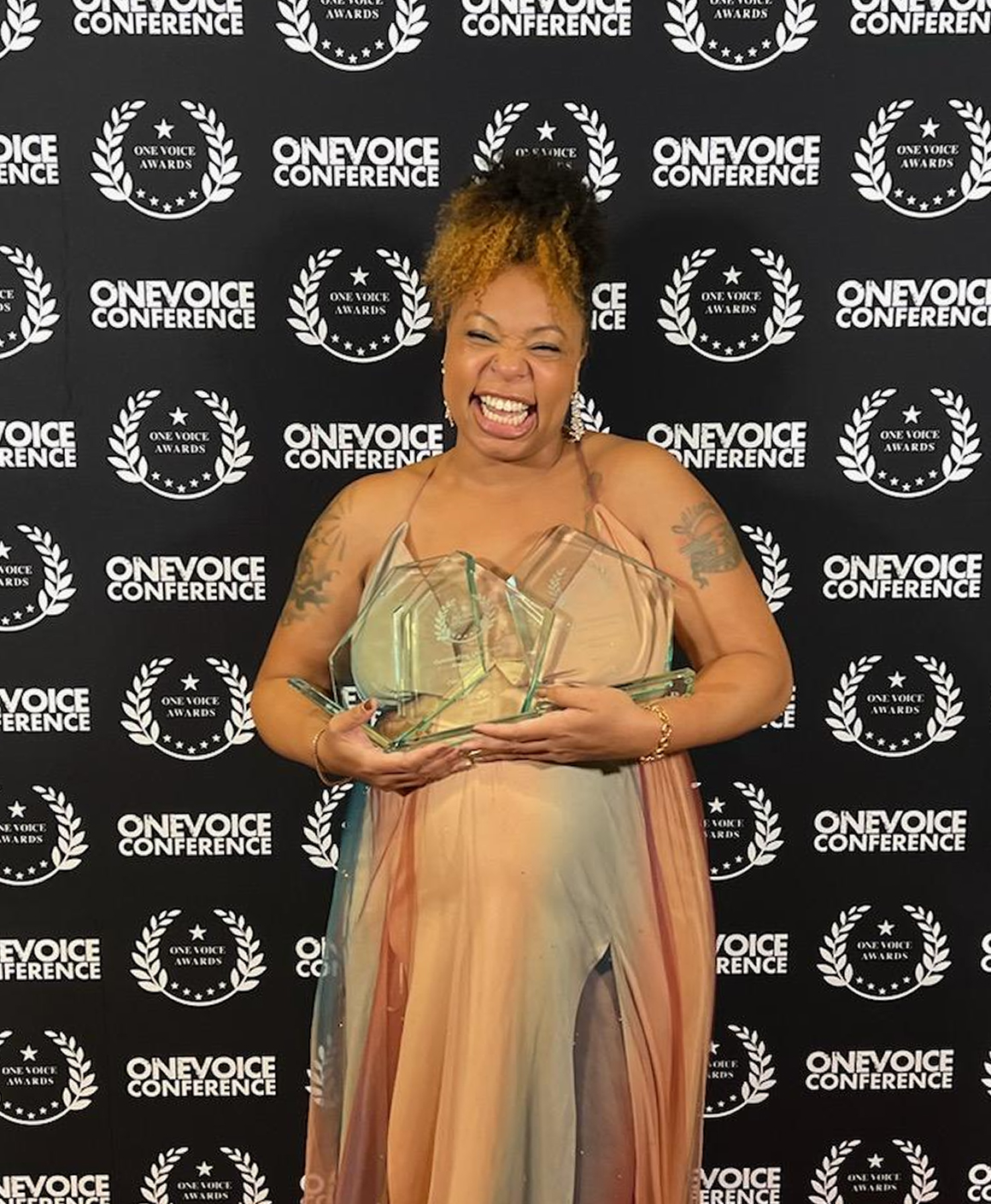 A person in a colorful dress smiles while holding two glass awards in front of a backdrop with "One Voice Conference" and award logos, celebrating achievements and inspiring others on how to get better at storytelling.