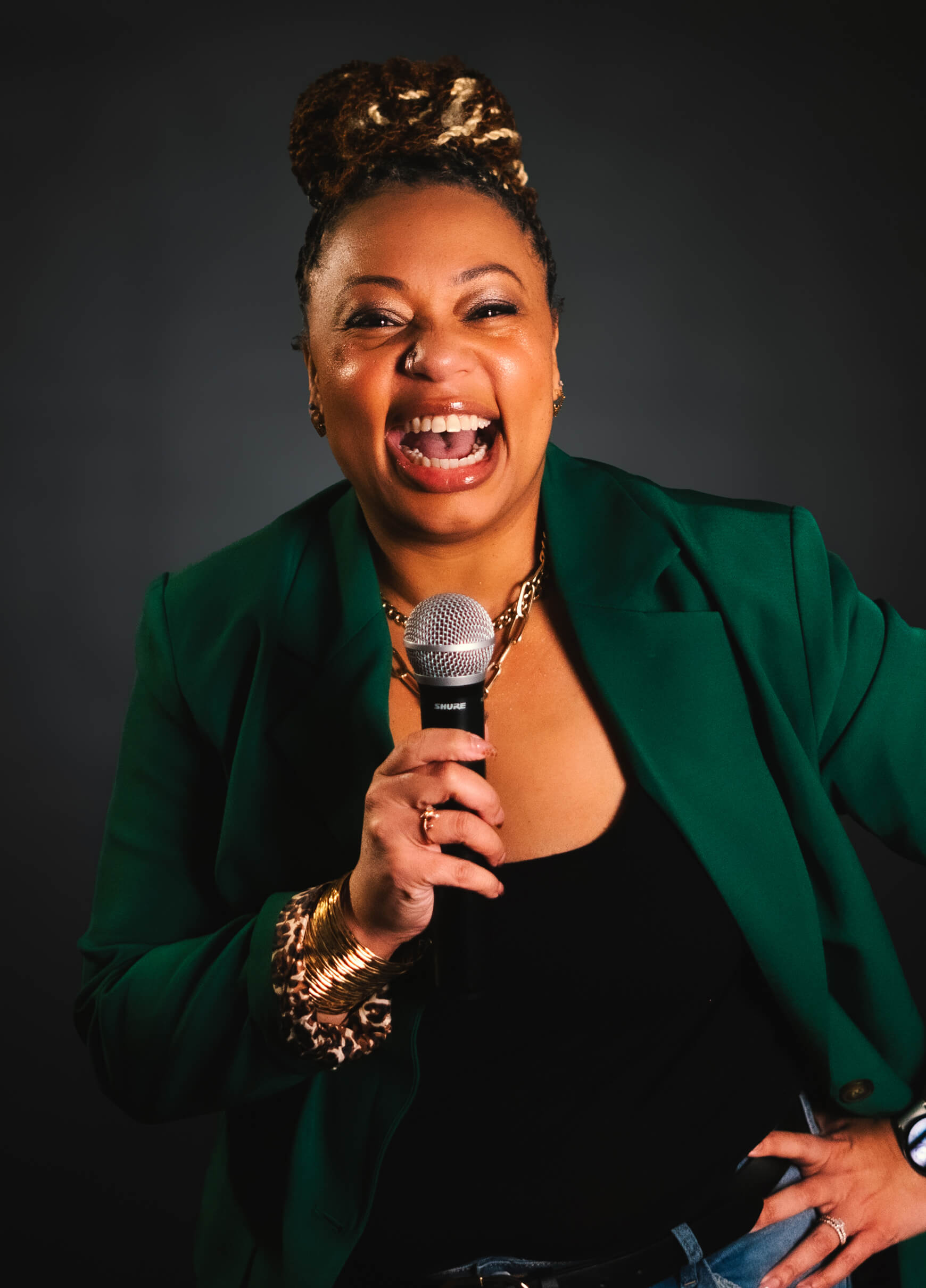 A person with braided hair in a green blazer holds a microphone and smiles widely against a dark background.