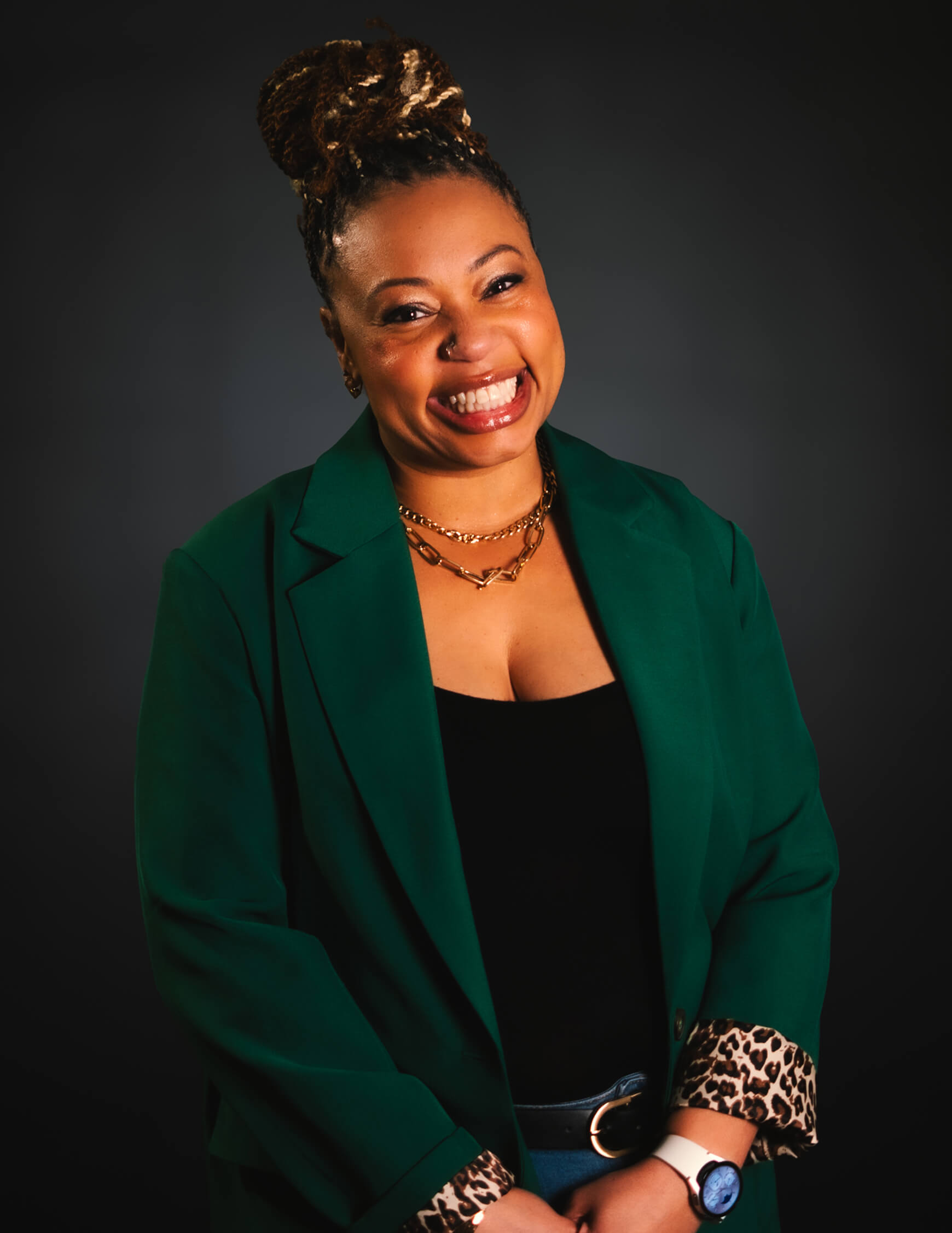 Smiling woman with braided hair wearing a green blazer over a black top, gold necklace, and a watch, standing against a dark background.