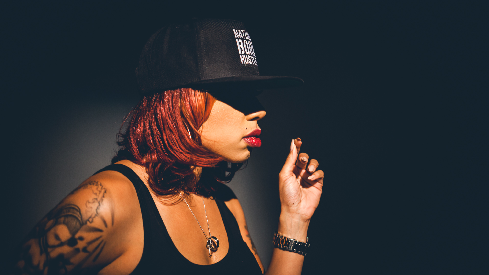 Woman with red hair, tattoo, and bright lipstick wears a black cap and tank top, holding her hand up near her face against a dark background.