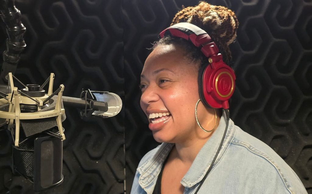 A person wearing red headphones and a denim jacket speaks or sings into a microphone in a soundproof recording booth.