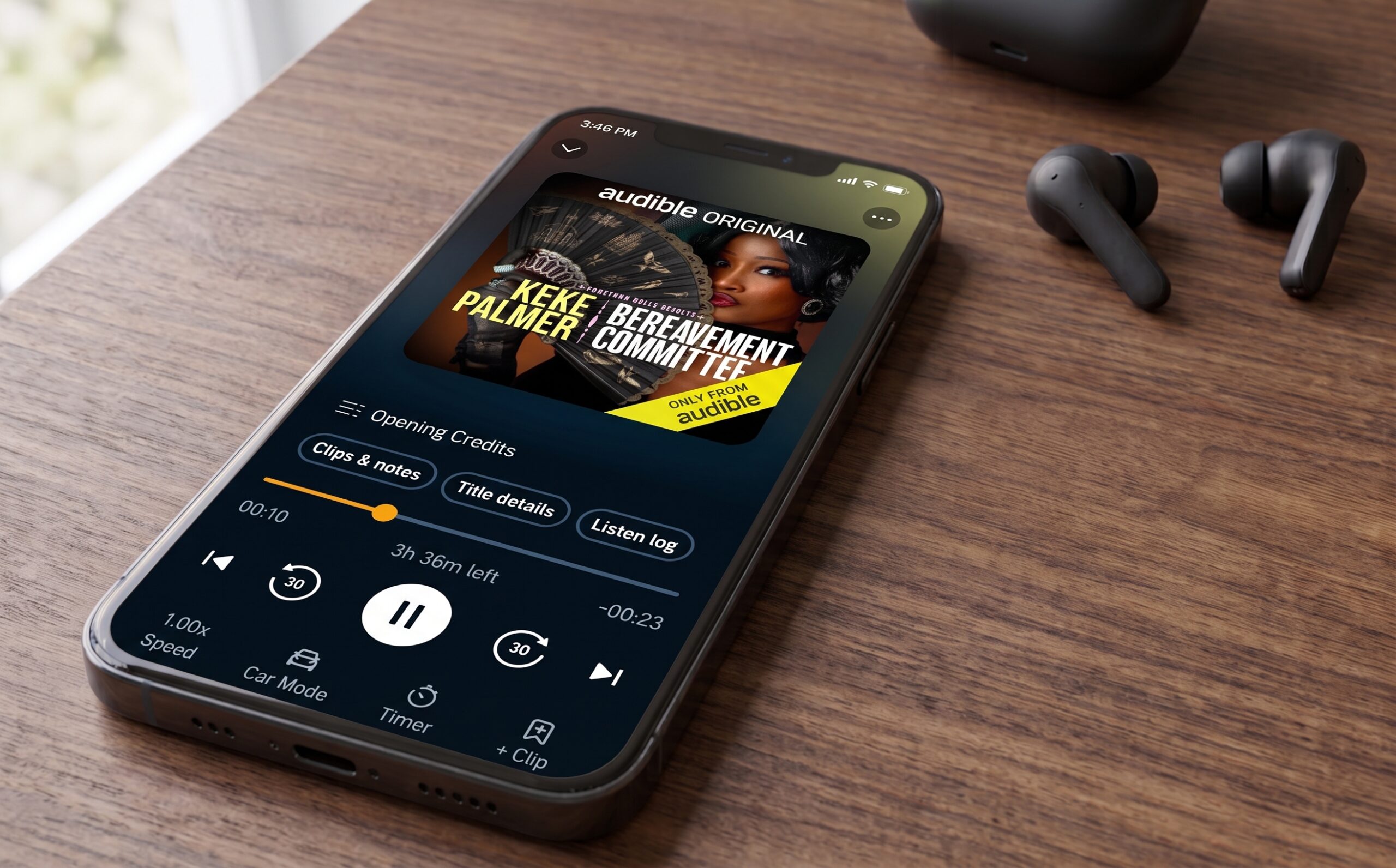 A smartphone on a wooden table displays the Audible app playing “Keke Palmer: The Backup” audiobook. Wireless earbuds are placed next to the phone.
